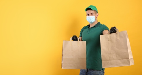 Photo of Courier in medical mask holding paper bags with takeaway food on yellow background, space for text. Delivery service during quarantine due to Covid-19 outbreak