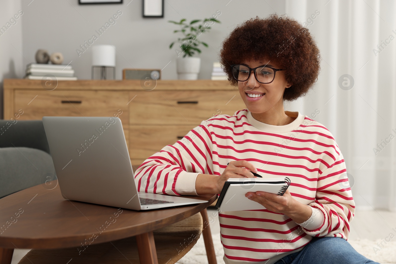 Photo of Beautiful young woman using laptop and writing in notebook at wooden coffee table in room