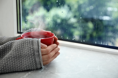 Image of Woman with cup of hot drink near window on rainy day, closeup