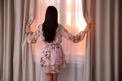 Young woman wearing floral print dress near window at home, view from back