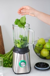Photo of Woman adding spinach leaves into blender with ingredients for smoothie at table indoors, closeup