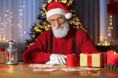 Photo of Santa Claus at his workplace. Letters, cup of beverage and gift boxes on table in room with Christmas tree