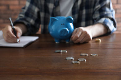 Photo of Man with piggy bank counting money at wooden table, focus on scattered coins