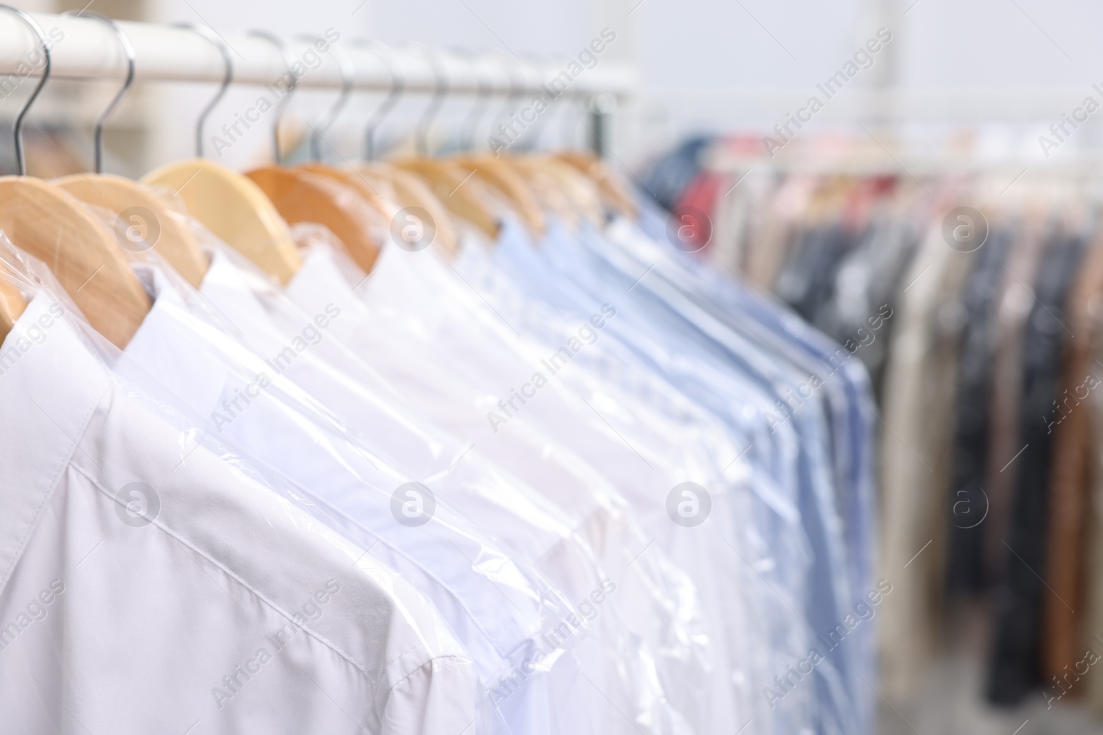 Photo of Dry-cleaning service. Many different clothes in plastic bags hanging on rack indoors, closeup