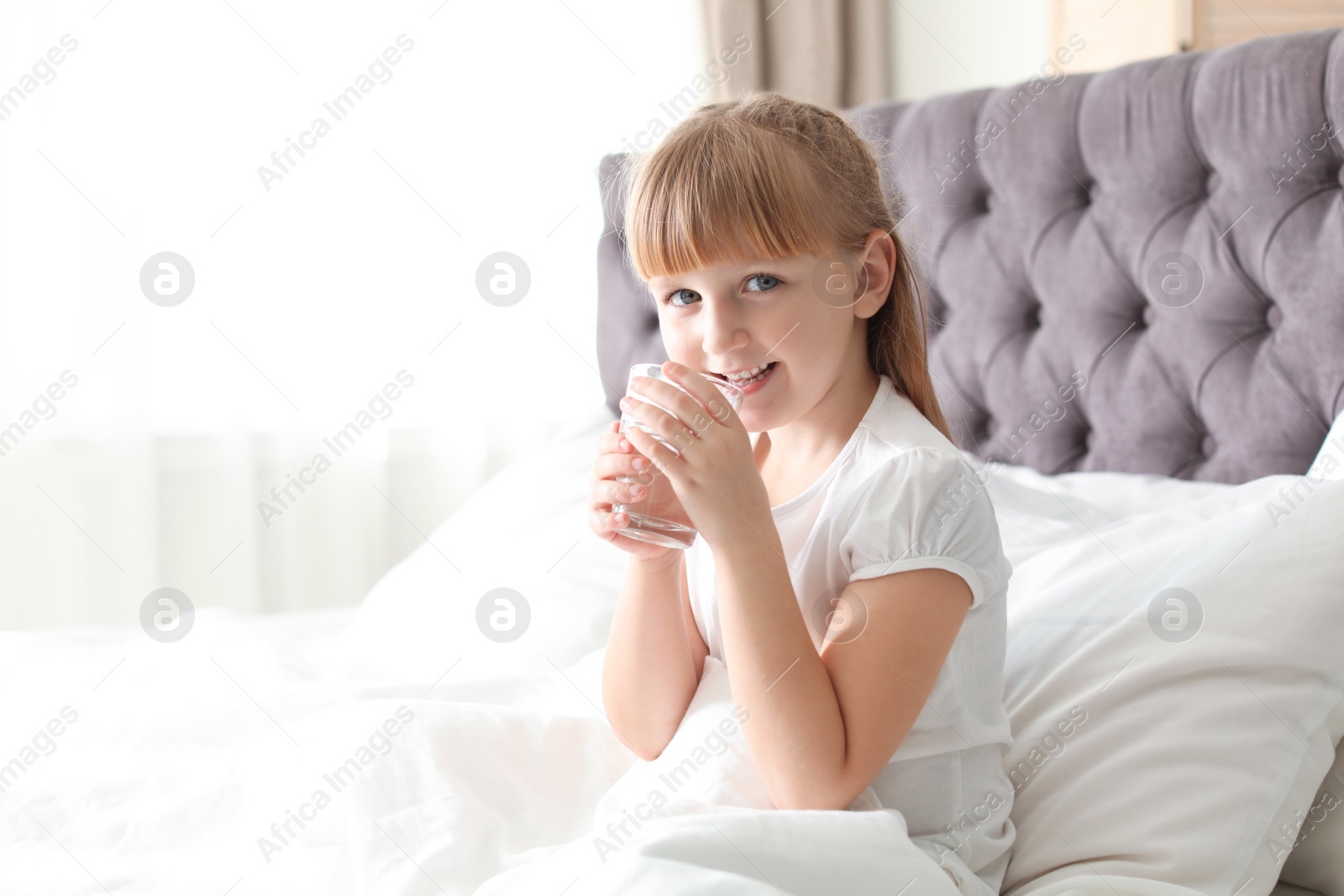 Photo of Cute little girl with glass of fresh water sitting in bed at home. Space for text