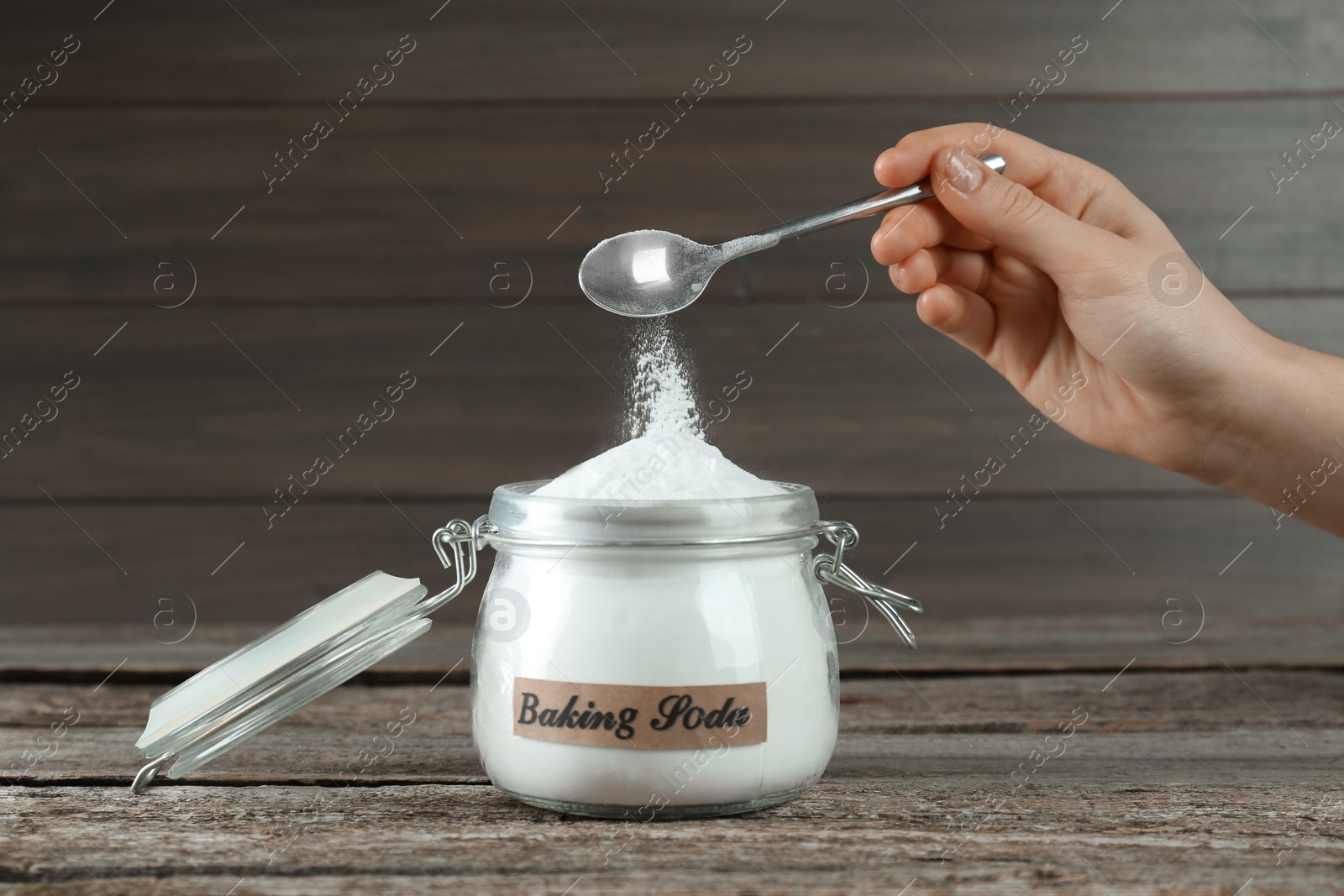 Photo of Woman pouring baking soda from spoon into jar at wooden table, closeup