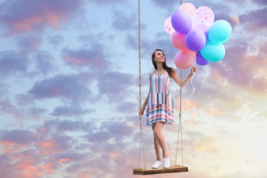 Image of Dream world. Young woman with bright balloons on swing in sunset sky