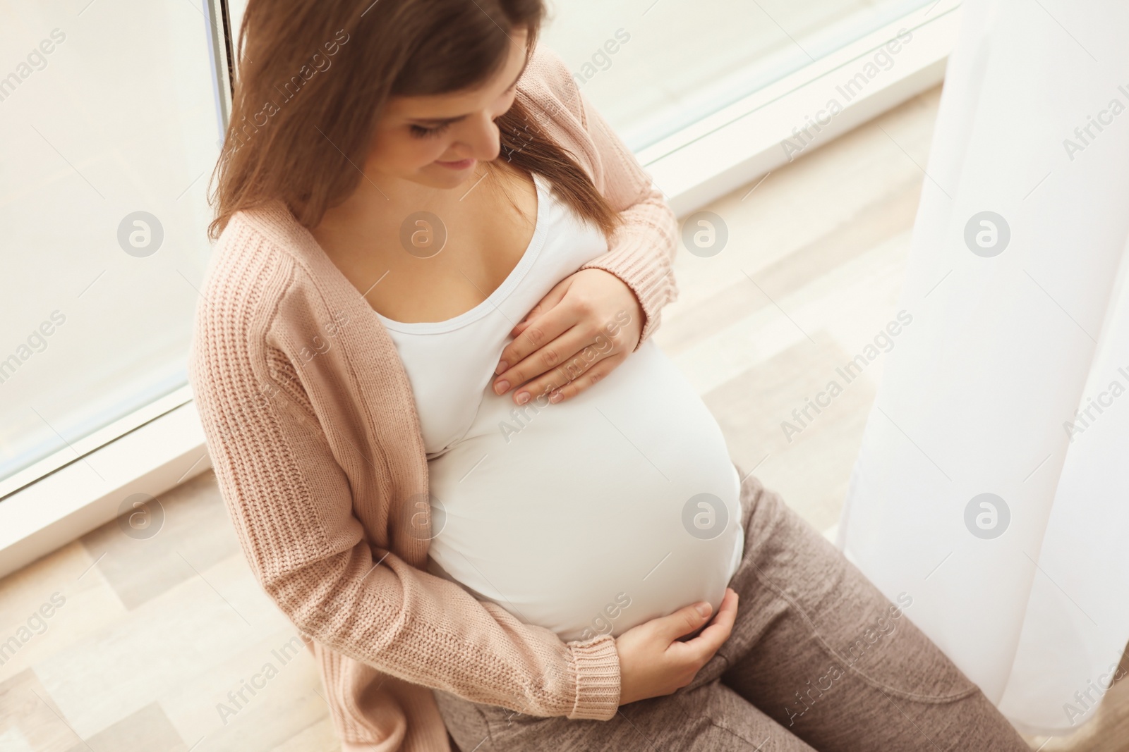 Photo of Young pregnant woman sitting on window sill at home