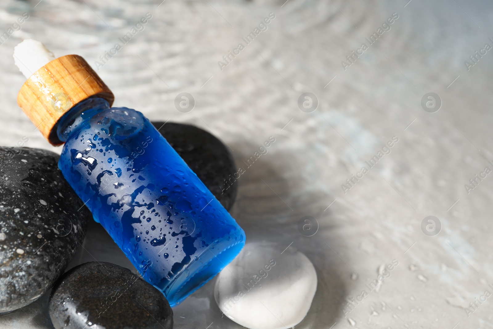 Photo of Bottle of face serum and spa stones in water on light background, closeup. Space for text