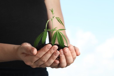 Photo of Woman holding green hemp plant in soil against blue sky, closeup