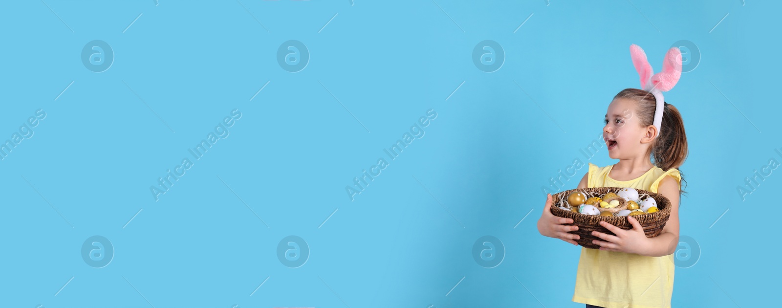 Photo of Excited little girl with bunny ears holding wicker basket full of Easter eggs on light blue background. Space for text