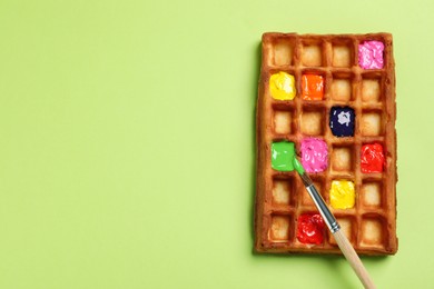 Photo of Waffle with different paints and brush on light green background, top view. Space for text