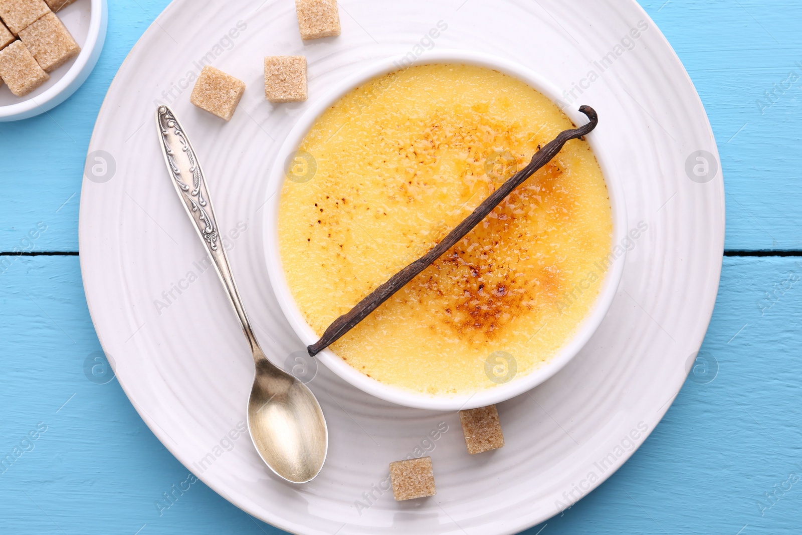 Photo of Delicious creme brulee in bowl, vanilla pod, sugar cubes and spoon on light blue wooden table, top view