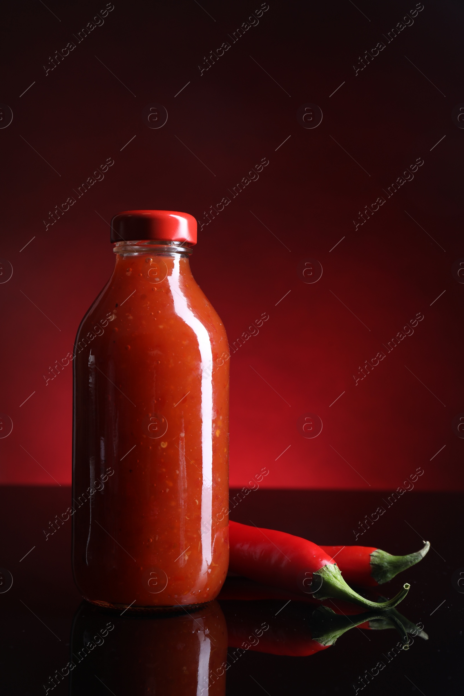 Photo of Spicy chili sauce in bottle and peppers against dark background, space for text