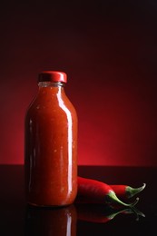 Photo of Spicy chili sauce in bottle and peppers against dark background, space for text