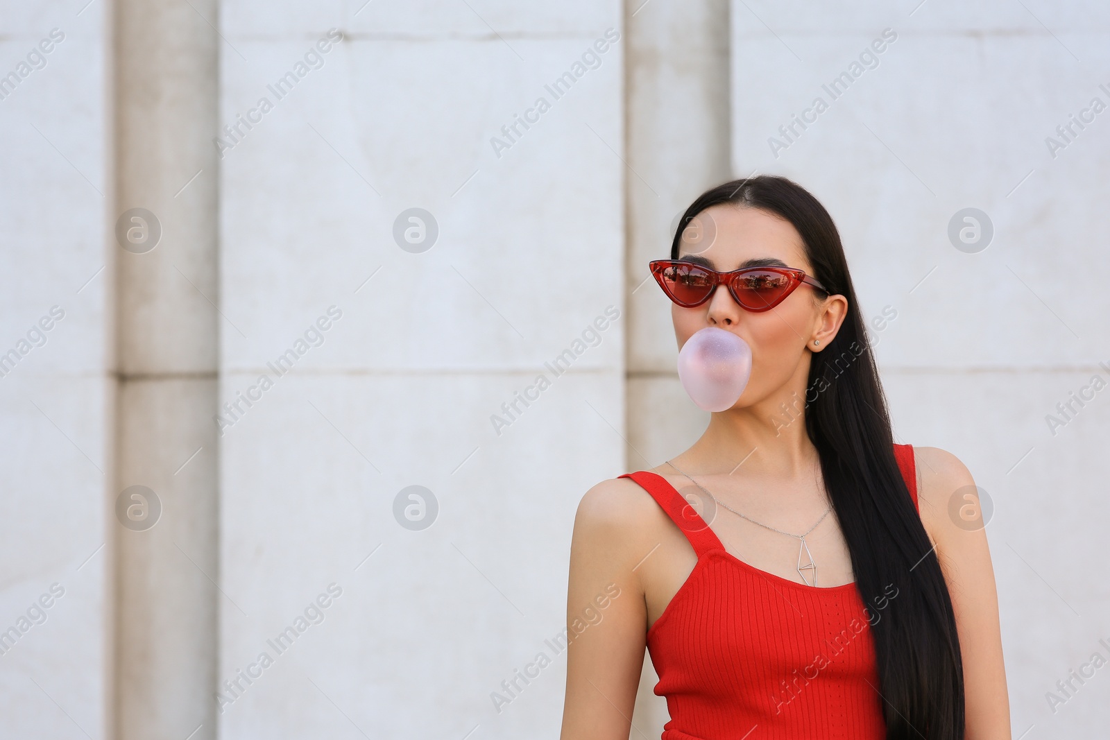 Photo of Beautiful woman in stylish sunglasses blowing gum near wall outdoors, space for text