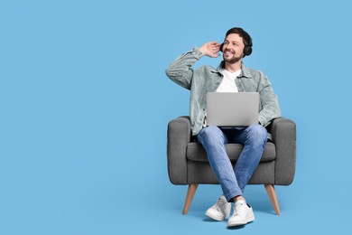 Happy man with laptop and headphones sitting in armchair on light blue background, space for text
