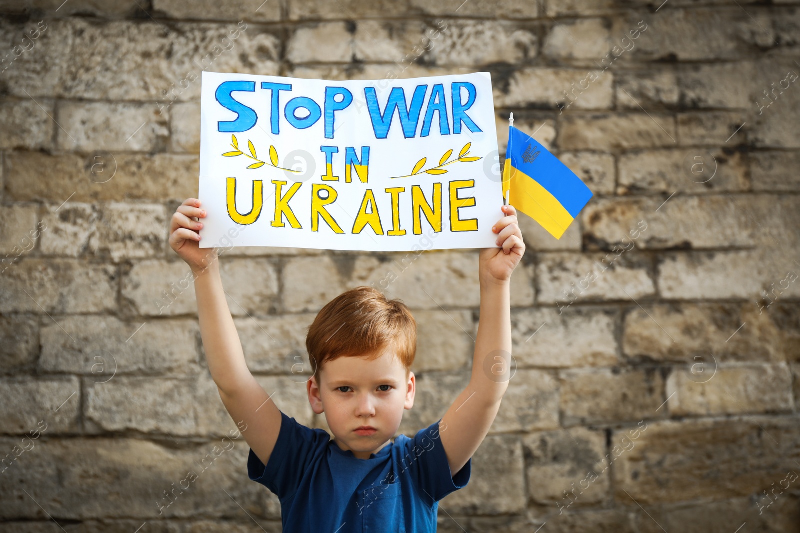 Photo of Sad boy holding poster Stop War In Ukraine and national flag against brick wall outdoors