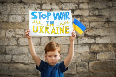 Photo of Sad boy holding poster Stop War In Ukraine and national flag against brick wall outdoors