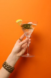 Photo of Woman holding martini glass of refreshing cocktail with lemon slice and rosemary on orange background, closeup