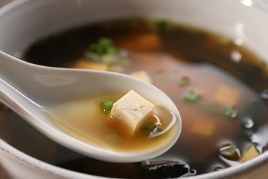 Delicious miso soup with tofu in spoon above bowl, closeup