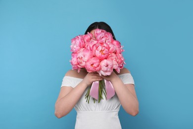 Photo of Young woman covering her face with bouquet of pink peonies on light blue background