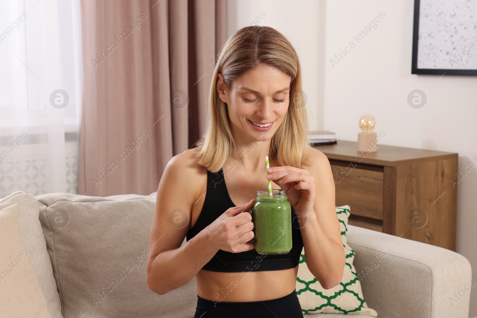 Photo of Young woman in sportswear with mason jar of fresh smoothie at home