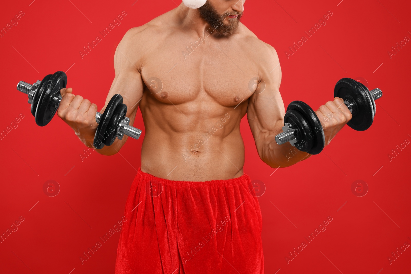 Photo of Muscular young man in Santa hat with dumbbells on red background, closeup