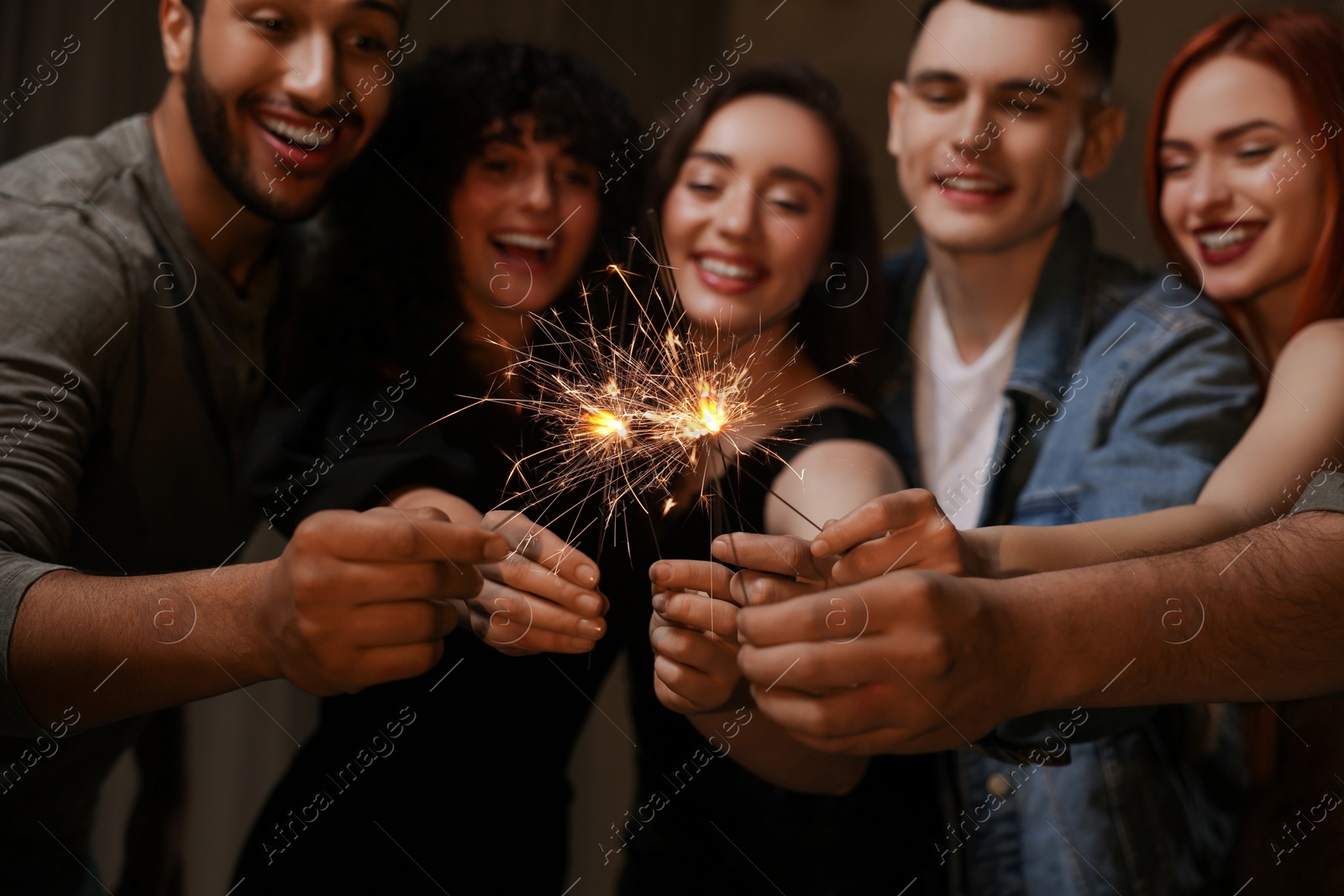 Photo of Happy friends with sparklers celebrating birthday indoors, focus on hands