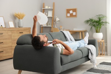 Photo of Man waving white hand fan to cool himself on sofa at home