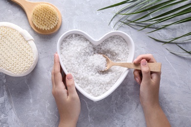 Photo of Woman holding heart shaped plate with sea salt for spa scrubbing procedure at grey marble table, top view