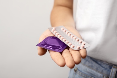 Young woman holding birth control pills and condom on light grey background, closeup. Safe sex concept