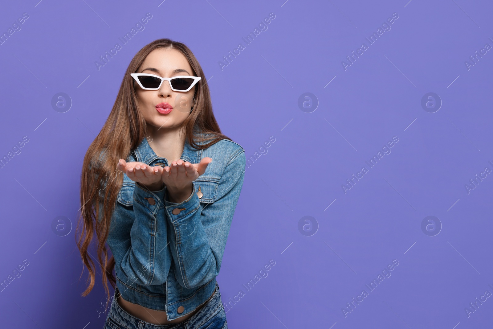 Photo of Beautiful young woman with sunglasses blowing kiss on purple background, space for text