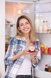 Photo of Young attractive woman eating tasty yogurt near fridge