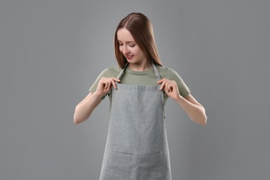 Photo of Beautiful young woman wearing kitchen apron on grey background. Mockup for design