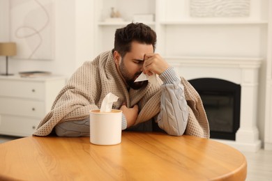 Photo of Sick man wrapped in blanket with tissue blowing nose at wooden table indoors. Cold symptoms