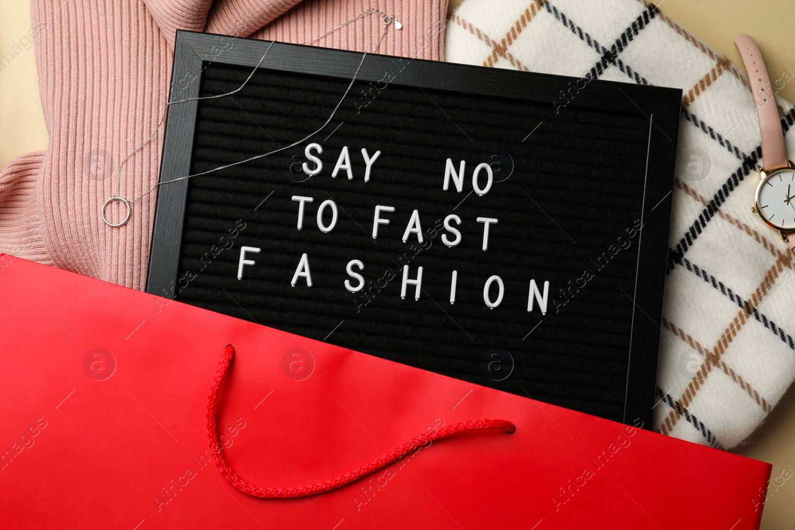 Photo of Black letter board with phrase SAY NO TO FAST FASHION, clothes and wristwatch in red shopping bag, top view