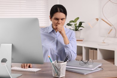Photo of Woman coughing while using computer at table in office. Cold symptoms