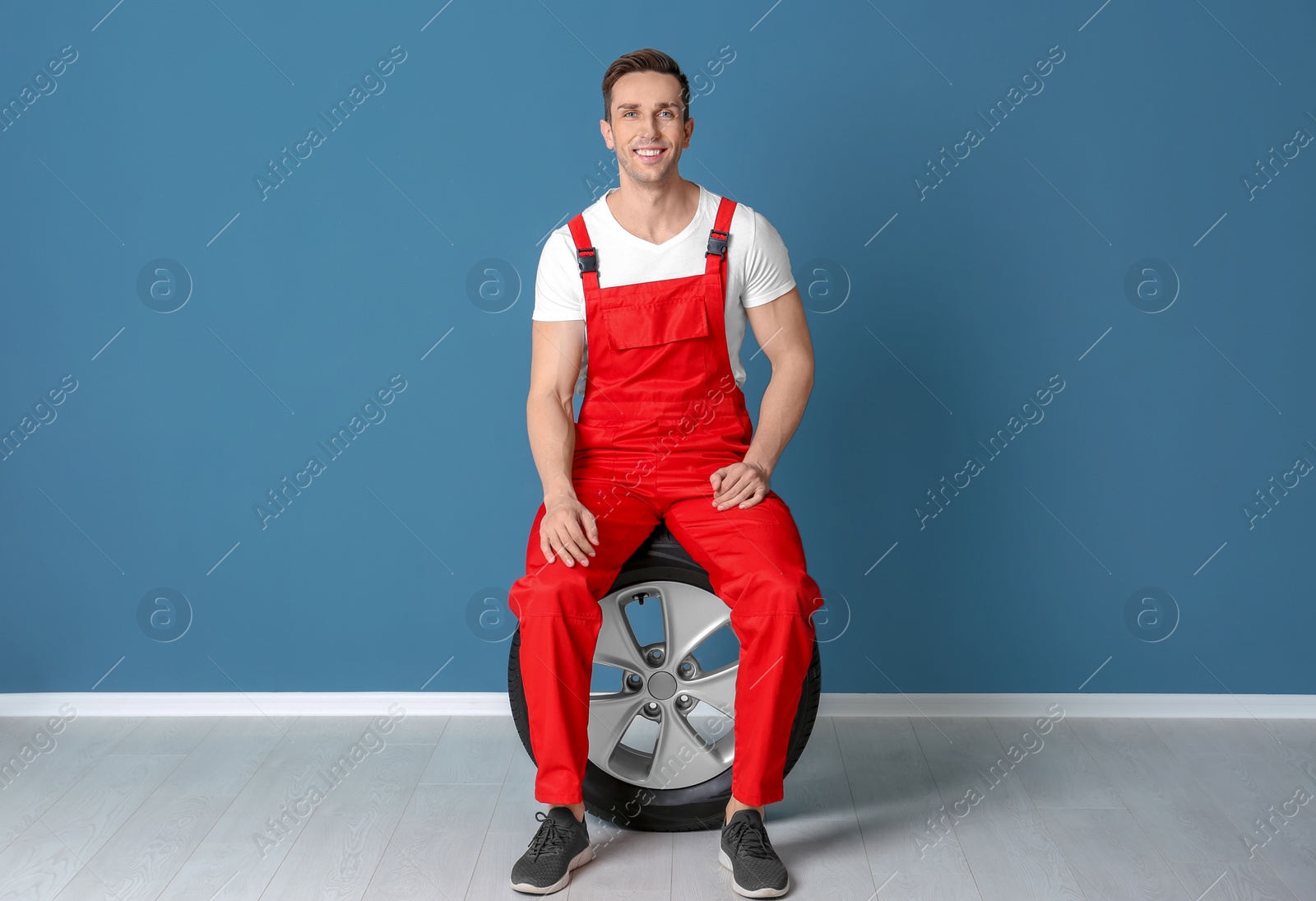 Photo of Young mechanic in uniform with car tire near color wall