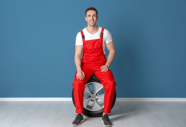 Young mechanic in uniform with car tire near color wall