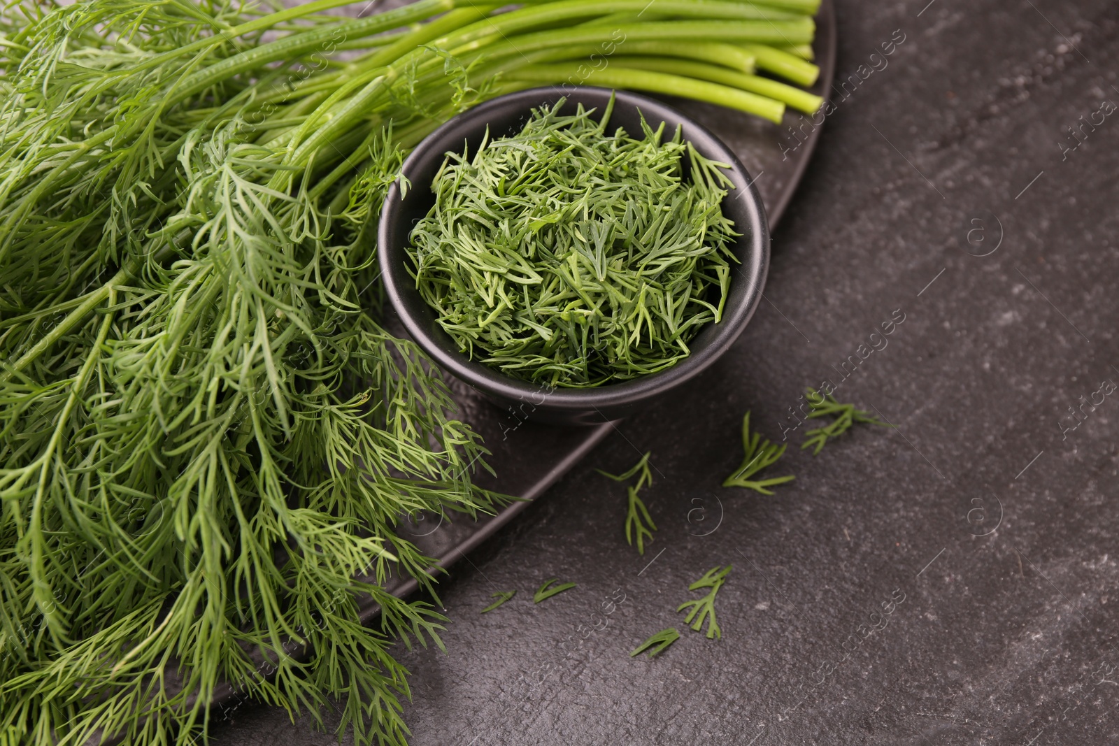 Photo of Sprigs of fresh dill and bowl with cut one on dark textured table, closeup