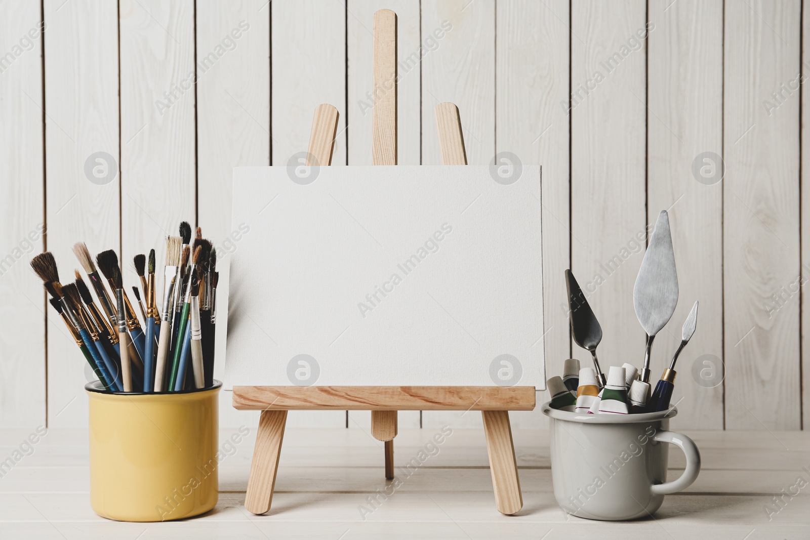 Photo of Easel with blank canvas, paints and brushes on white wooden table