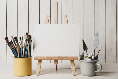 Easel with blank canvas, paints and brushes on white wooden table