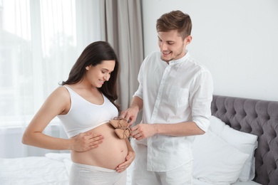 Young husband touching his pregnant wife's tummy with cute little shoes in bedroom