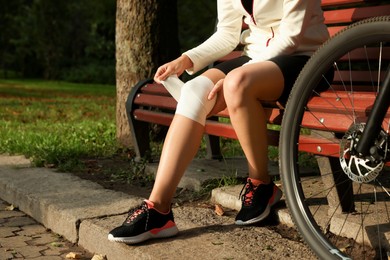 Photo of Young woman applying bandage onto her knee on wooden bench outdoors, closeup