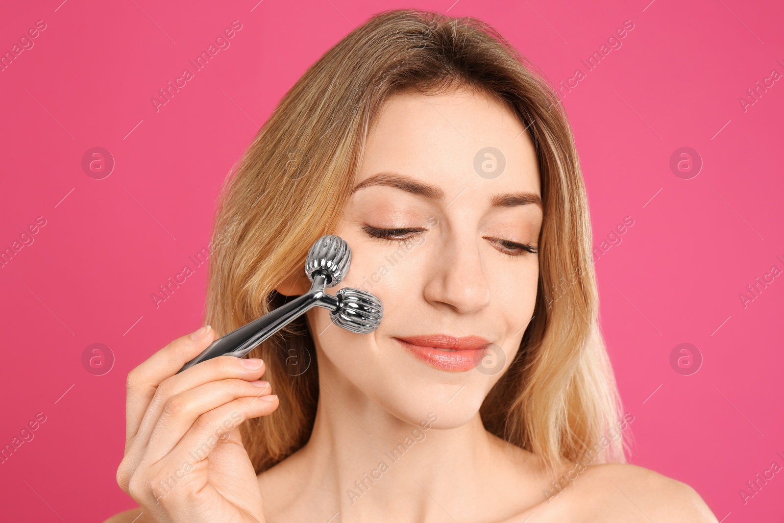 Photo of Young woman using metal face roller on pink background