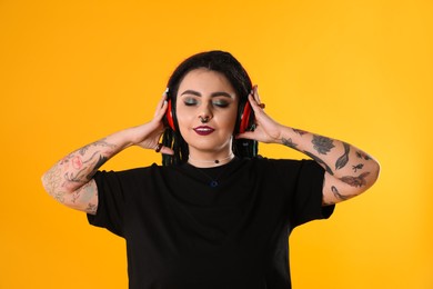Beautiful young woman with tattoos on arms, nose piercing and dreadlocks listening music against yellow background