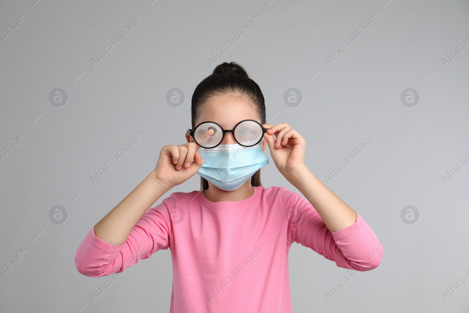 Photo of Little girl wiping foggy glasses caused by wearing medical face mask on grey background. Protective measure during coronavirus pandemic