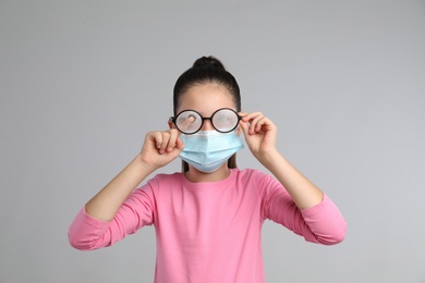 Photo of Little girl wiping foggy glasses caused by wearing medical face mask on grey background. Protective measure during coronavirus pandemic