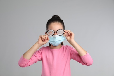 Little girl wiping foggy glasses caused by wearing medical face mask on grey background. Protective measure during coronavirus pandemic